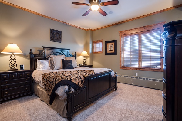 Your cozy bedroom features a plush queen bed with elegant linens, warm bedside lamps, and a ceiling fan for your comfort. Large windows invite natural light into your serene mountain retreat.