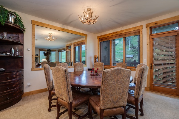 Gather around this elegant dining table, where rustic antler chandeliers cast a warm glow and large windows frame peaceful views, making every meal feel special in your mountain retreat.