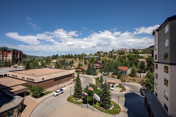 Mountain resort setting with nearby residential buildings, lush evergreens, and clear summer skies. The property is located in a scenic hillside community with convenient access to local amenities.