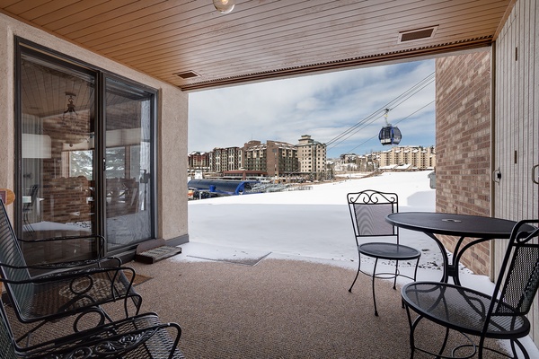 A covered patio with seating overlooks a snowy ski resort base area, offering views of slopes, gondola access, and surrounding village buildings amid winter scenery.