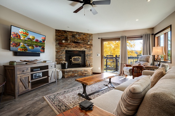 Unwind in your cozy living room featuring a stone fireplace, plush seating, and balcony access with serene views—perfect for fall evenings and movie nights.