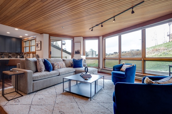 Sink into plush seating and let natural light flood your space through floor-to-ceiling windows framing mountain views. Your cozy retreat features warm wood ceilings and stylish furnishings perfect for unwinding after adventure.