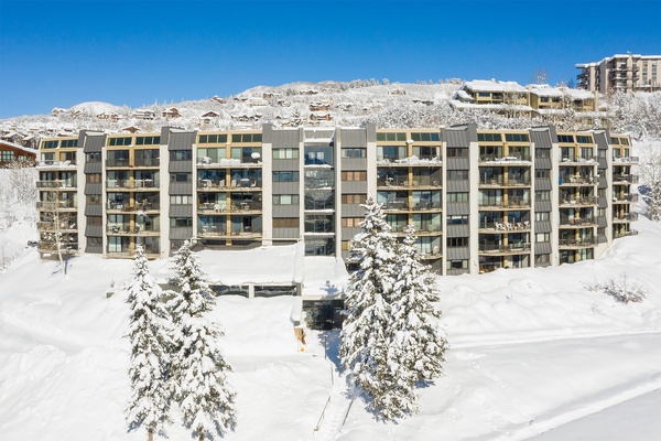 This modern ski resort building sits amid a stunning winter landscape, its multiple balconies overlooking snow-blanketed slopes and frosted evergreens under crisp blue skies.