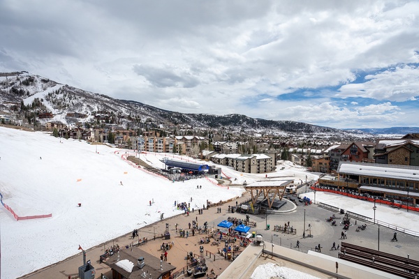 Aerial view of a mountain resort village nestled against snow-covered slopes, featuring ski facilities, lodges, and a bustling base area with shops and restaurants.