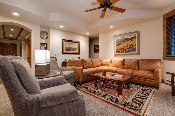 Sink into plush seating and unwind in your spacious living room, where a cozy sectional and armchairs invite relaxation beneath a gently circulating ceiling fan and warm ambient lighting.