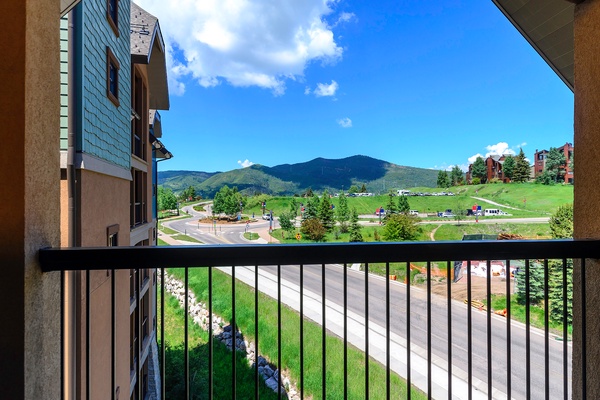 Panoramic mountain views stretch across rolling green hills and forests, framed by a balcony overlooking the surrounding resort area and distant peaks.
