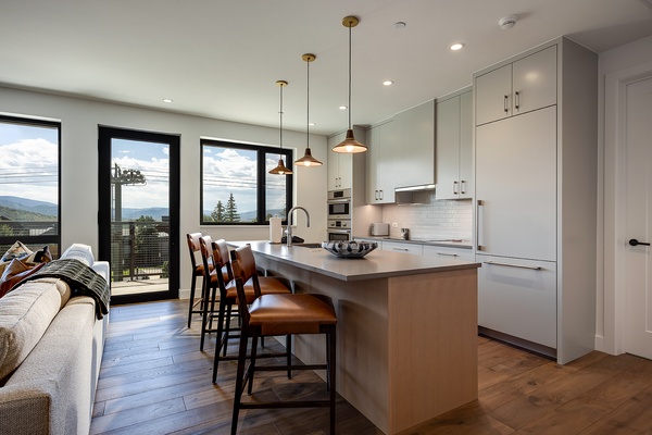 Step into your open-concept kitchen with sleek white cabinetry, a spacious island for gathering, and warm pendant lighting that sets the mood. Mountains frame your mornings through floor-to-ceiling windows.