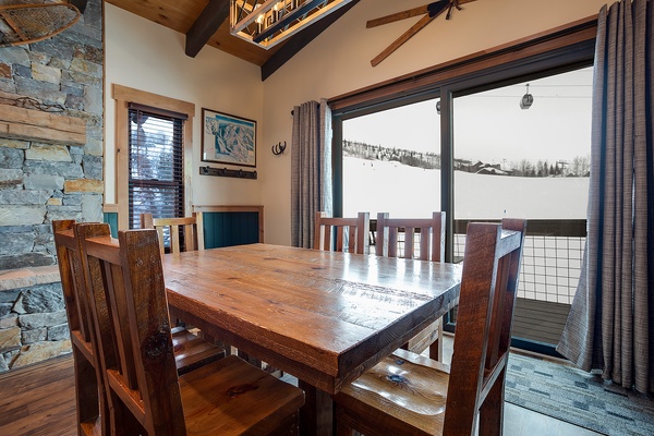Gather around the handcrafted wooden table in your dining area, where floor-to-ceiling windows frame breathtaking ski slope views. The vaulted ceiling, stone fireplace, and mountain scenery create an unforgettable alpine retreat ambiance.
