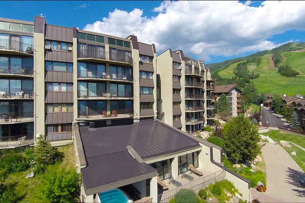 Aerial view captures the property nestled in a mountainside community with ski slopes, summer greenery, and neighboring buildings defining this alpine vacation destination.