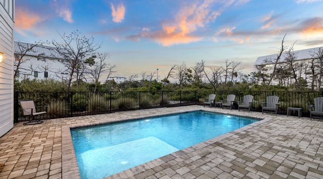 The property features a private pool surrounded by comfortable loungers, set against a backdrop of natural coastal vegetation and colorful evening skies.