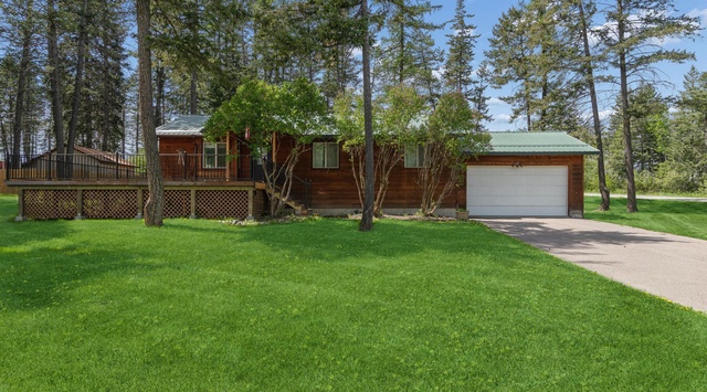 A charming cedar cabin nestled among towering pines on a spacious property. The home features a wrap-around deck, attached garage, and lush green lawn—surrounded by natural beauty and peaceful forest views.