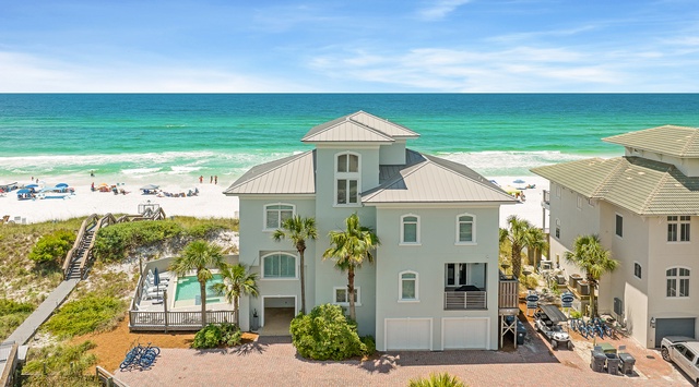 A stunning beachfront property features emerald-green Gulf waters, private beach access, and coastal landscaping with palm trees. The three-story home sits steps from pristine white sand and vibrant turquoise waves.