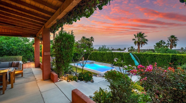 A stunning sunset paints the sky above the property's pool area, framed by lush gardens, hedges, and palm trees in the surrounding landscape.