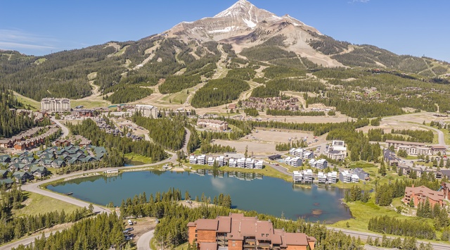 An aerial view captures the property nestled in a picturesque mountain resort setting, surrounded by alpine forests, ski slopes, and a tranquil lake reflecting the stunning peaks beyond.