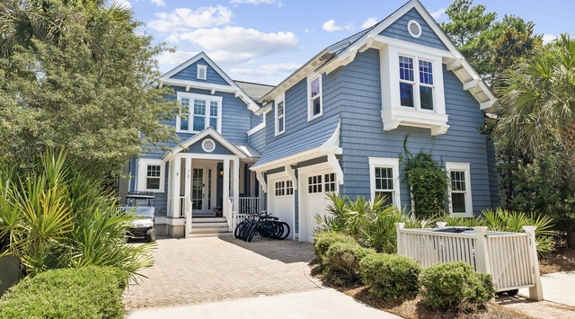 Charming coastal-style home with blue siding, white trim, and welcoming front porch, surrounded by lush tropical landscaping and palms.