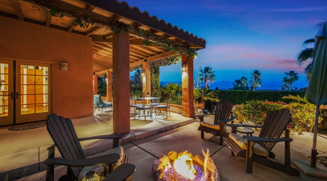 An elegant covered patio overlooks lush gardens and tropical palms, where you can gather around the glowing fire pit beneath a stunning twilight sky—perfect for relaxing evenings outdoors.