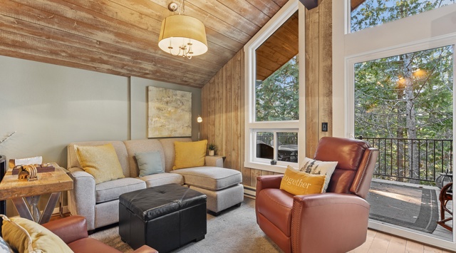 Relax in your spacious living room featuring vaulted wood ceilings, leather seating, and floor-to-ceiling windows overlooking lush treetops.