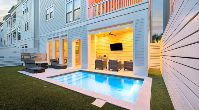 A stunning three-story coastal property features a private pool surrounded by lush turf, with a covered outdoor lounge beneath a second-floor balcony. Multiple seating areas invite you to unwind in style at sunset.