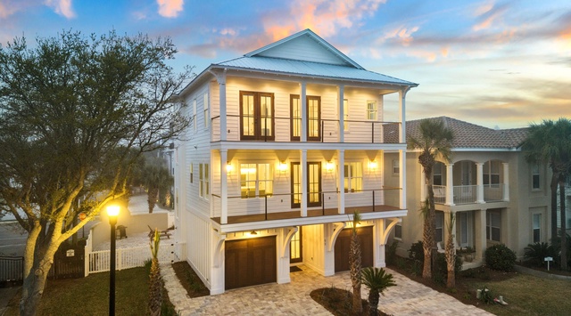 Stunning coastal home showcases elegant three-story design with warm lighting and beautiful landscaping in a peaceful neighborhood setting.