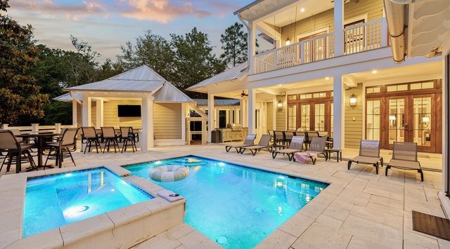 A beautiful vacation home featuring a private pool and spa surrounded by comfortable loungers, a covered outdoor dining pavilion with TV, and elegant two-story architecture nestled among towering pines.