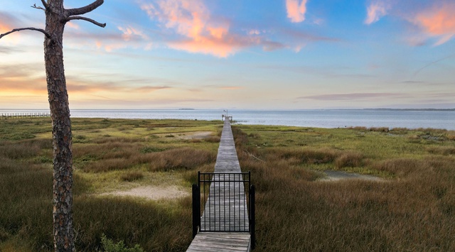A wooden boardwalk stretches across coastal marshland toward the water, surrounded by natural grasses and golden evening light painting the sky.