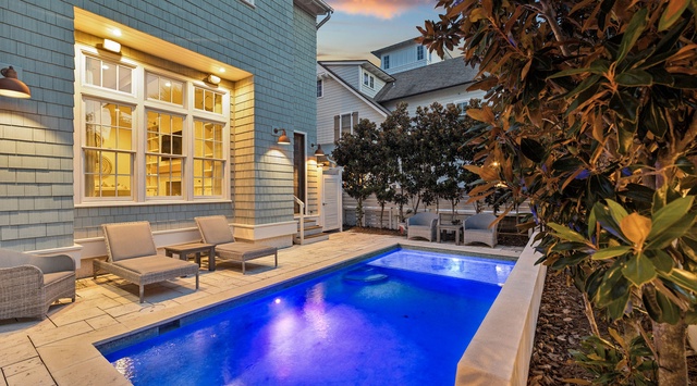 The property features a private illuminated pool framed by manicured landscaping and neighboring coastal architecture, creating an inviting evening atmosphere.