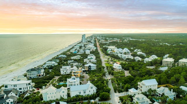Aerial view of coastal community with pristine beaches, tree-lined streets, and charming homes nestled between ocean and forest at sunset.