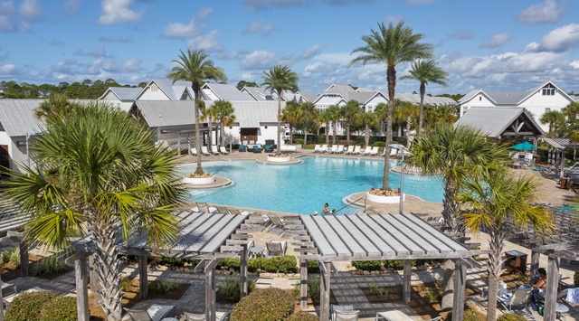 Resort community with central pool area surrounded by white cottage-style buildings and tropical palm landscaping under partly cloudy skies.