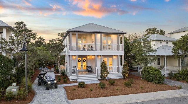 A charming two-story coastal cottage with wraparound porches and welcoming yellow doors, nestled in a peaceful residential neighborhood with paver walkways and golf cart access.