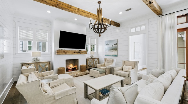 Sink into plush seating by the fireplace as natural light fills this elegant living space, where rustic wood beams meet coastal charm for your perfect retreat.