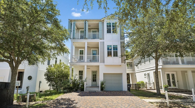 A welcoming three-story coastal home nestled among shade trees with stacked balconies and a brick paver driveway leading to the garage.