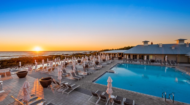 A stunning beachfront pool deck bathed in golden sunset light, with lounge chairs and umbrellas awaiting your arrival. The property's distinctive architecture frames the serene ocean horizon perfectly.
