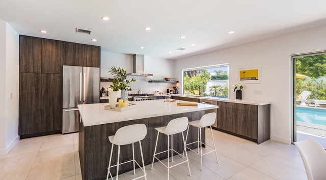 Imagine crafting your morning coffee in this sleek, modern kitchen with its rich wood cabinetry and bright white countertops. Natural light pours through wide windows, while your sparkling pool beckons just beyond the glass doors.