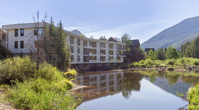Nestled beside a tranquil pond with mountain peaks rising behind, the property sits within a lush natural setting surrounded by evergreens and vibrant greenery.