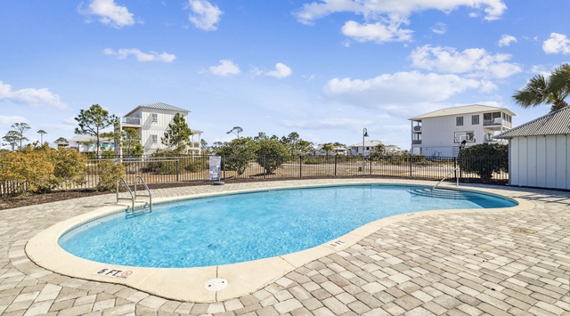 A sparkling community pool awaits, surrounded by coastal pines and brick pavers, with nearby beach homes completing this inviting neighborhood retreat for your coastal escape.