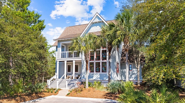 Charming coastal home with tropical landscaping and spacious porches, nestled among lush trees and palms in a serene residential setting.