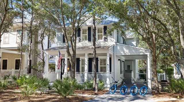 A charming two-story white beach home nestled among coastal trees, featuring a welcoming front porch with bicycles ready for exploring the neighborhood.