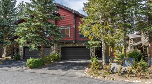 A charming red mountain-style property nestled among evergreen and deciduous trees, with stone accents and a convenient two-car garage, located in a peaceful neighborhood setting.