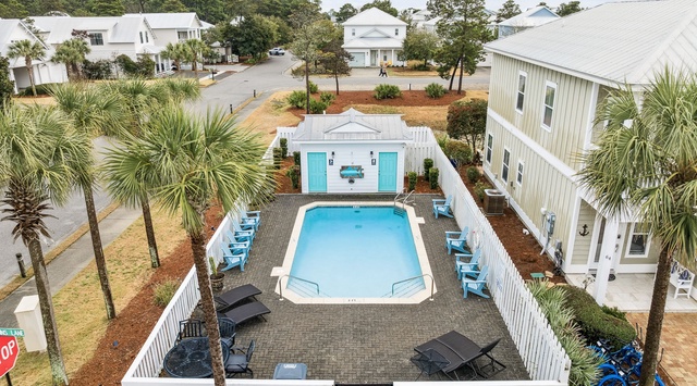 An aerial view showcases the property's inviting pool area, complete with lounge chairs and a small pool house, all nestled within a charming coastal neighborhood lined with palm trees and nearby homes.