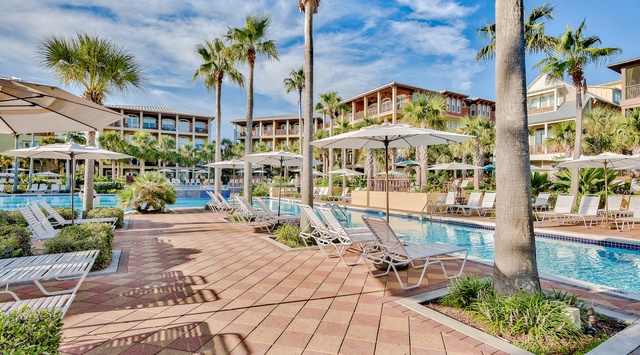 Tropical resort setting with swimming pools surrounded by palm trees and multi-story buildings under a bright blue sky.
