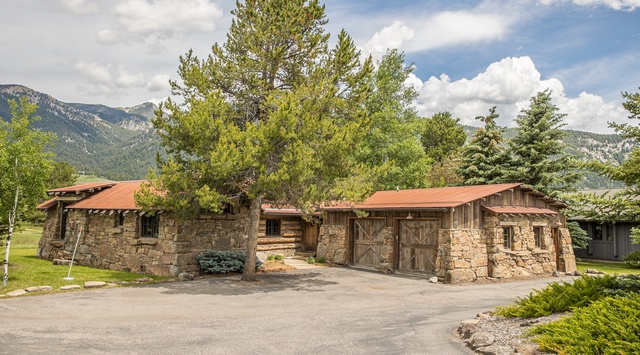 A stunning mountain retreat showcasing rustic stone and timber architecture, surrounded by lush evergreens and majestic peaks under a dramatic sky.