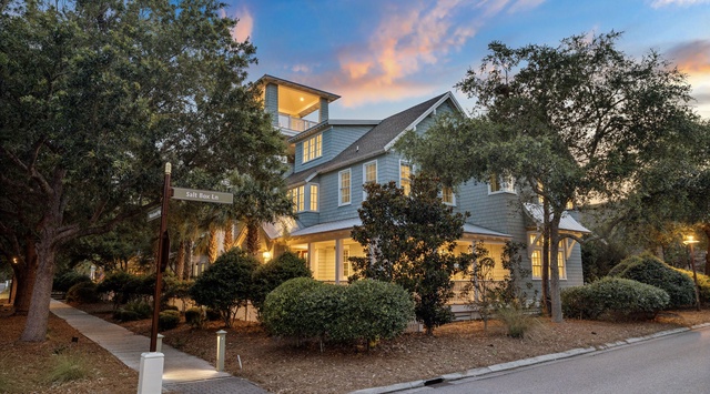 A charming coastal property nestled among mature trees, with warm lighting glowing through windows as sunset colors paint the evening sky.