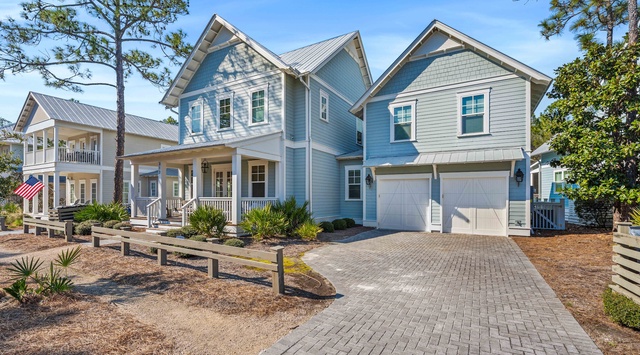 Charming coastal-style home with a welcoming front porch and convenient two-car garage, nestled among mature pines in a peaceful neighborhood.