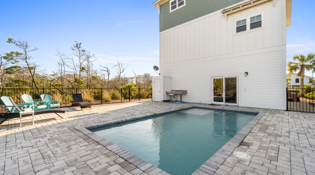 A modern coastal retreat featuring a private pool surrounded by paver stone decking. The fenced backyard includes colorful lounge chairs, a grill station, and palm tree-dotted landscaping.
