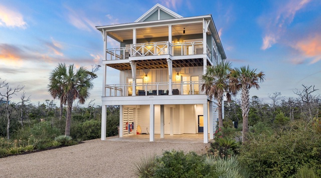 A striking coastal retreat with multiple levels and expansive decks, nestled among native palms and natural vegetation, captured at twilight as warm lighting illuminates the home's elegant architecture.