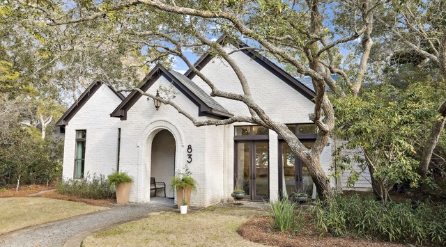 This charming painted brick home features an arched entrance and mature oak trees creating natural shade over the welcoming front yard.