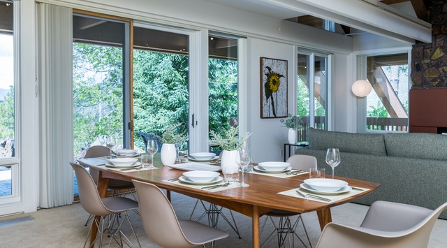 Gather around the warm walnut table set for your group, with sunlight streaming through glass doors that open to lush greenery—your perfect spot for memorable meals together.