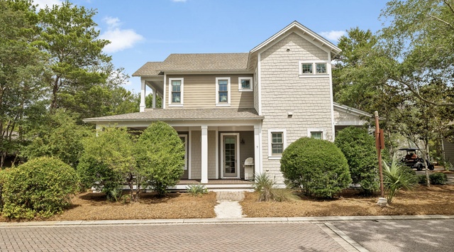 A charming two-story vacation home with modern farmhouse styling, nestled among mature trees in a peaceful residential setting with professional landscaping and covered porch entry.