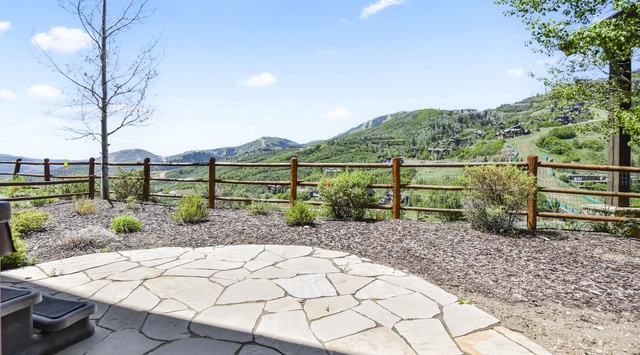 Expansive stone patio overlooking rolling mountain slopes and valleys. A peaceful outdoor space with rustic wooden fencing and natural landscaping, ideal for relaxing and enjoying scenic mountain vistas.
