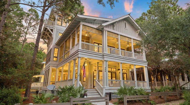 A graceful coastal home nestled among towering pines, featuring wraparound porches and warm interior lighting that glows through oversized windows beneath a painted dusk sky.